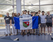 Tony Shi, au centre, tenant le drapeau ténois au côté de ses équipiers et de ses coachs au Centre aquatique de Yellowknife peu de temps avant de s'envoler vers les Jeux d'été du Canada 2025. — Ollie Williams/Team NT
