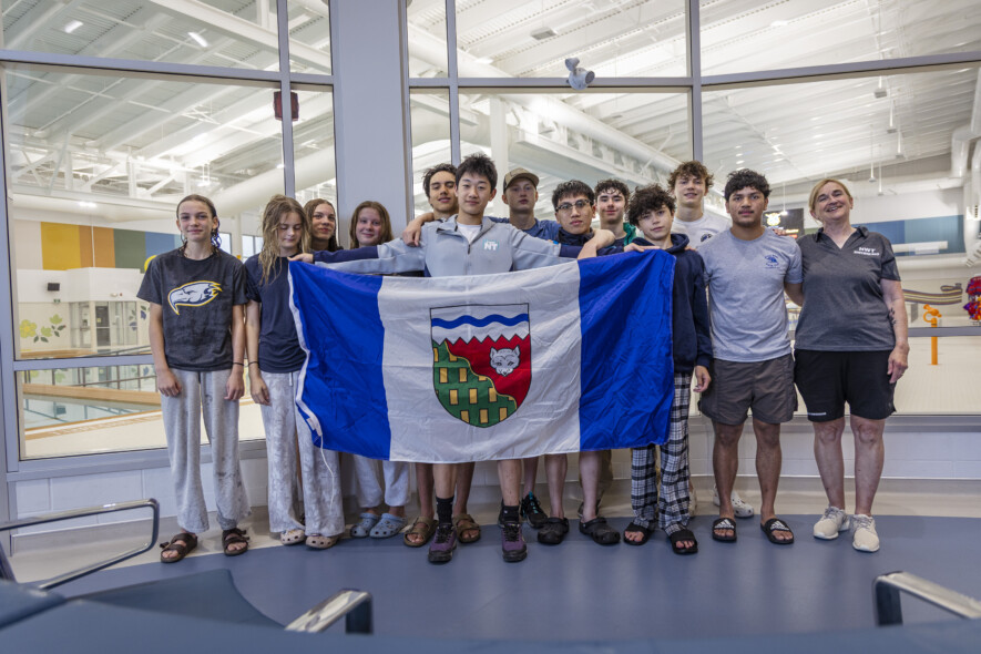 Tony Shi, au centre, tenant le drapeau ténois au côté de ses équipiers et de ses coachs au Centre aquatique de Yellowknife peu de temps avant de s'envoler vers les Jeux d'été du Canada 2025. — Ollie Williams/Team NT