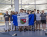 Tony Shi, au centre, tenant le drapeau ténois au côté de ses équipiers et de ses coachs au Centre aquatique de Yellowknife peu de temps avant de s'envoler vers les Jeux d'été du Canada 2025. — Ollie Williams/Team NT