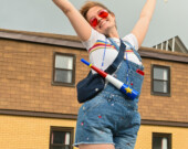 Un membre du Tintamarre de la Fierté pose pour une photo, tenant un drapeau trans dans une main et un bruiteur portant un drapeau acadien autour du cou.  &mdash; PHOTO - Shannon Nickerson